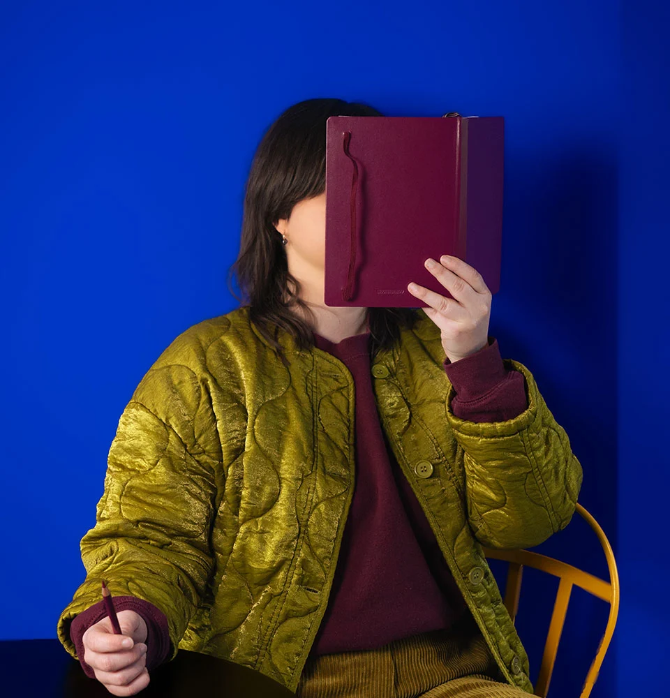 bleue-brut-dans-son-carnet- Bleue Brut tient son carnet devant son visage, elle a un crayon dans la main.